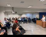  Imagem de um auditório com pessoas sentadas em cadeiras e ao centro uma mulher.