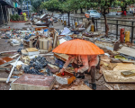 Imagem de uma pessoa, abaixada, segurando um guarda chuva, na calçada, no meio dos destroços causados pela enchente no RS. 