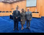 Trêsq pessoas posando para foto dentro de um plenário. Uma mulher ao centro, ladeada por dois homens.