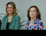 Foto de duas mulheres claras à frente de uma parede branca. A da esquerda está sorrindo e usa óculos e blazer verde. A da direita tem semblante sereno e usa vestido com estampa geométrica colorida.