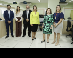 Foto de seis pessoas, sendo dois homens e quatro mulheres, de pé, sorrindo e posando para a câmera. Elas estão em um ambiente claro com divisórias nas laterais e placas de identificação próximas ao teto.
