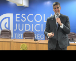 Foto de um homem claro, usando terno cinza, segurando um microfone com a mão esquerda e uma caneta com a direita, ao mesmo tempo em que sorri para a câmera. Ele está posicionado à direta da foto. Atrás dele, vê-se uma parede azul com a logomarca branca da Escola Judicial e uma mesa amadeirada com quatro cadeiras marrons. À frente da mesa, vê-se brasão dourado da República do Brasil.