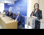 Foto de uma mulher em pé, atrás de um púlpito transparente, à direita, usando óculos e vestido escuro, e falando ao microfone, que segura com a mão direita. À esquerda, 4 pessoas sentadas a uma mesa de madeira, tendo às suas costas parede azul com a inscrição ESCOLA JUDICIAL em letras brancas.