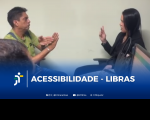 Foto mostra, à esquerda, um homem claro de cabelos escuros, usando camisa verde e se comunicando por meio de sinais com as mãos. À direita, vê-se mulher clara de cabelos escuros, compridos e lisos, usando blusa e blazer pretos. Ela conversa em LIBRAS com o homem. Ambos estão sentados em frente a uma parede bege. Abaixo, tarja azul degradê, onde se vê a logomarca do TRT-16 e o texto branco ACESSIBILIDADE - LIBRAS. Abaixo, estão os endereços do TRT-16 nas redes sociais.