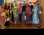 A imagem mostra uma foto de grupo em frente a um painel informativo sobre a "Preservação da Memória". As pessoas estão vestidAs com roupas casuais e alguns estão sorrindo para a câmera. O fundo é um espaço interno com piso de madeira.