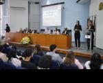 Foto mostra à direita mulher clara com roupa preta, atrás de um pulpito falando ao microfone. A sua direita, posiciona-se mulher com roupa preta, interprete de libras. A direita desta, mesa de honra com a presença de seis pessoas sentadas.