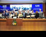 Foto mostra um grupo formado por homens e mulheres, em pé, atrás de uma bancada. Ao fundo, um painel eletrônico ligado com o destaque para a logomarca da ALEMA - Assembléia Legislativa do Maranhão