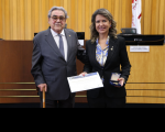 Foto mostra um homem e uma mulher segurando um certificado e uma medalha. Na parte do fundo, imagem de uma parede amaderada.