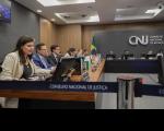 Foto de pessoas sentadas a uma mesa em forma de L, tendo à sua frente computadores e microfones. Ao fundo paredes amadeiradas e cinza escuro, onde aparece a logomarca do CNJ em metal prateado.