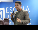 Foto de um homem claro e jovem, usando camisa e blazer caqui. Ele fala ao microfone, que segura com a mão direita. Atrás dele parede azul com inscrição branca. No canto inferior esquerdo, duas cadeiras marrons.