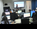 Foto de um homem claro, usando camisa cinza e dando aula para uma turma que usa computadores. Atrás dele, tela de projeção com apresentação de slides. O ambiente é uma sala de treinamento de informática com paredes brancas e azuis.