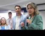 Foto de duas mulheres e dois homens. Em primeiro plano, à direita, mulher clara, de cabelos claros, usando blazer verde, fala ao microfone, que segura com a mão esquerda, e gesticula com a mão direita, olhando para a frente. Ao lado dela, as demais pessoas a observam falar.