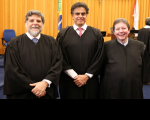 Foto de três homens claros, sorrindo e usando togas pretas de juiz. Atrás deles, parede amadeirada.