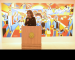Foto mostra uma mulher em pé, atrás de um púlpito de madeira. Ao fundo uma pintura colorida retrata figuras que representam trabalhadores
