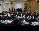 Foto mostra pessoas em apresentação no palco de um auditório. Outras pessoas estão sentadas nas cadeiras do auditório