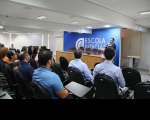 Foto mostra várias pessoas sentadas em um auditório. Ao fundo um homem, está em pé, diante do público.