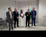 Foto mostra cinco pessoas em pé; Ao fundo uma parede branca com azulejos
