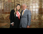 Foto mostra um homem e uma mulher. Ambos estão em pé. Ao fundo uma parede com azulejos marrons.