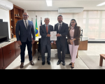 Foto mostra tres homens e uma mulher. Ambos estão em pé. Ao centro, os dois homens seguram uma folha de papel.