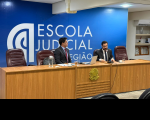 Foto mostra dois homens sentados à mesa de honra em uma auditório. Ao fundo o nome Escola Judicial