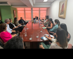 Foto mostra várias pessoas sentadas ao redor de uma mesa grande de reunião.