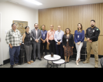 Foto mostra um grupo de pessoas em pé, dentro de uma sala. O grupo é formado por homens e mulheres.