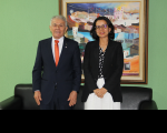 Foto mostra um homem e uma mulher, um ao lado do outro. Ambos estão em pé. Ao fundo uma parede na cor verde claro, e um quadro colorido posicionado na parede.