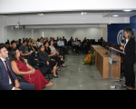 Foto mostra uma mulher em pé falando para uma platéia dentro de um auditório.