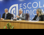 Foto mostra três pessoas sentadas. Ao fundo uma parede azul e o nome Escola Judicial