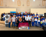 Foto mostra várias pessoas reunidas em um salão. Todos estão juntos em pé. São crianças, adolescentes e pessoas adultas reunidas.