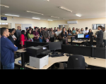 Foto mostra vários pessoas reunidas em pé, dentro de uma grande sala. 