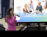 Foto mostra uma mulher usando um blaser na cor rosa. Ao fundo um telão projeto uma imagem com idosos reunidos. E a mensagem etarismo e ciclo da vida