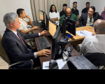 Foto mostra várias pessoas sentadas em volta de uma mesa. Sobre a mesa vários monitores de computador