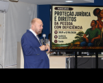 Foto mostra um homem falando ao microfone. Ao lado dele um painel com a informação: Proteção Jurídica e Direito da Pessoa com Deficiência