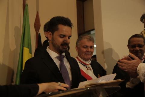 um homens de terno em pé, estando na tribuna lendo e o outros dois ao lado