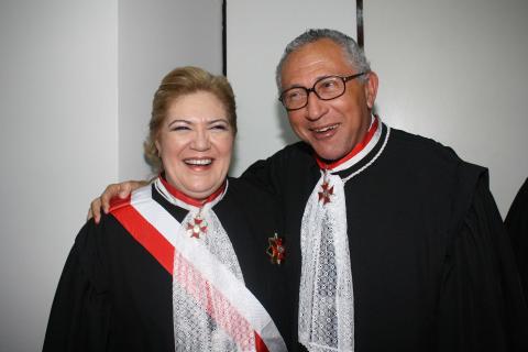 Mulher ao lado de um homem, ambos de toga pousando para foto. Ele pousa a mão sobre o ombro dela e ambos estão sorrindo para a foto