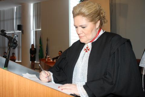 Mulher de toga em pé, assinando o ato de posse atrás de uma tribunal