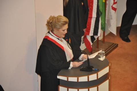 Mulher de toga em pé, atrás de uma tribuna segurando um papel e lendo o seu discurso