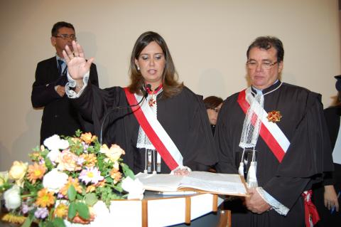 Mulher ao lado de um homem, ambos de toga, a mulher está com a mão estendida realizando juramento .