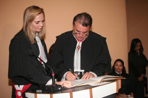 Mulher ao lado de um homem, ambos de toga, atrás de uma tribuna, o homem está assinando o ato de posse .