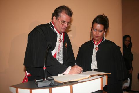 Mulher ao lado de um homem, ambos de toga, atrás de uma tribuna, o homem está assinando o ato de posse .