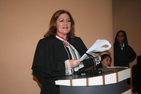 Mulher de toga em pé, atrás de uma tribuna, discursando.
