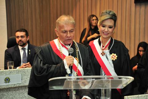 Mulher ao lado de um homem, ambos de toga, atrás de uma tribuna, o homem está com microfone .