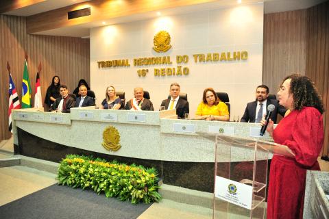 Mulher, em pé, em frente a um tribuna discursando, de vestido vermelho, atrás, mais para a direita, uma mesa com autoridades sentadas