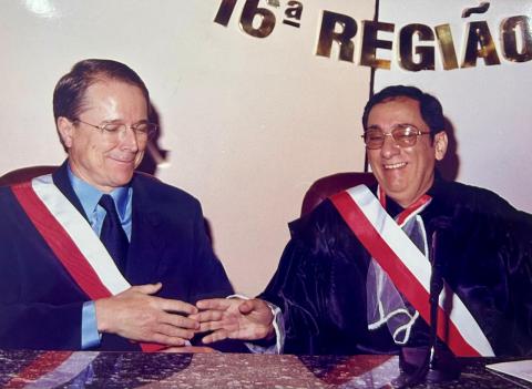 Dois homens de toga sentados atrás de uma mesa, um ao lado do outro, cumprimentando-se
