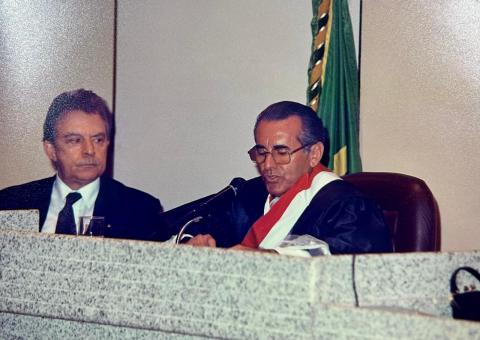 Homem de toga, sentado atrás de uma mesa,  falando em um microfone e ao lado um home de terno